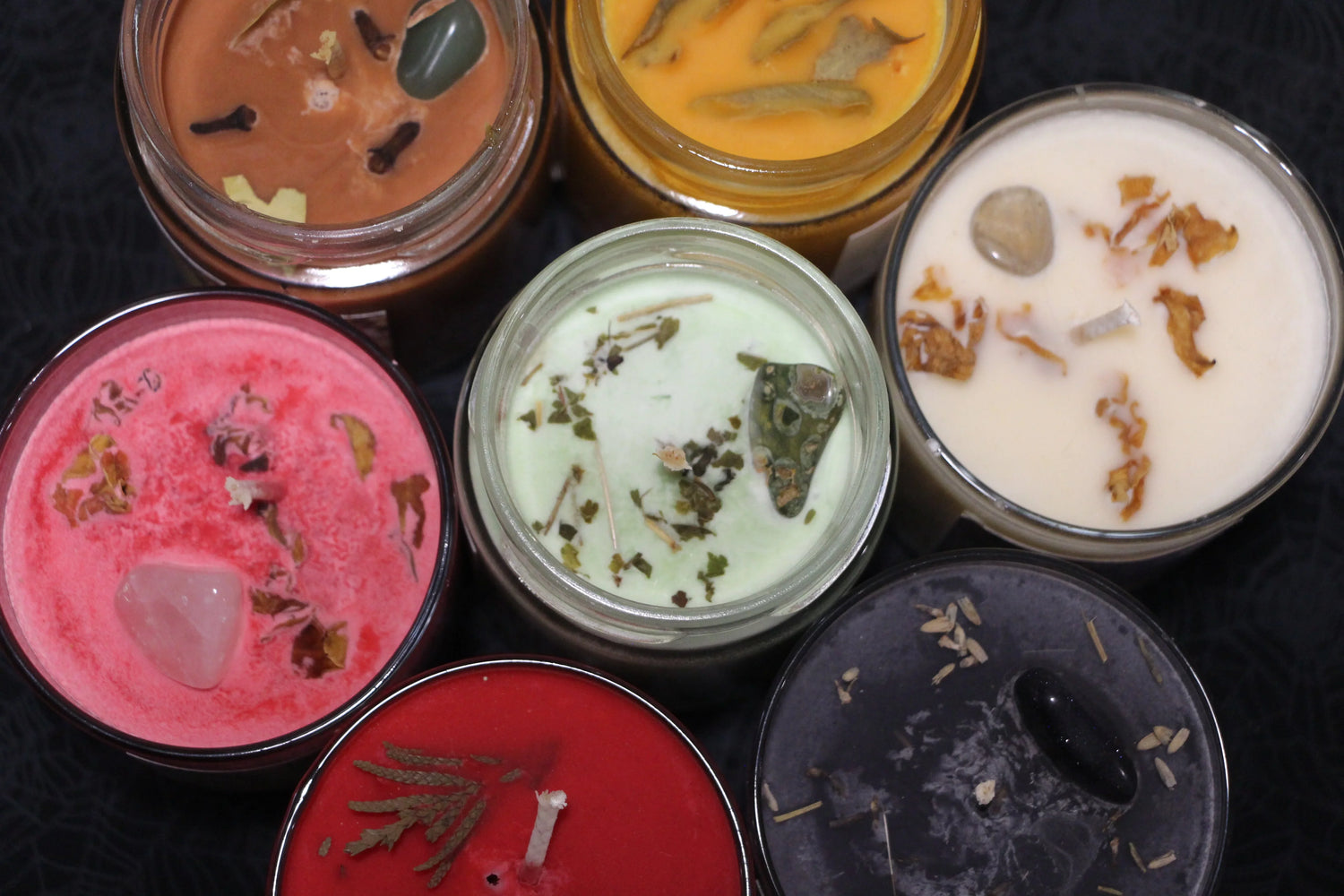 Top view of colorful handmade herbal and crystal-infused candles in glass jars on black surface